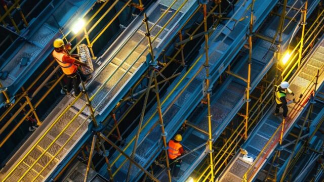 a dynamic construction site showcases skilled workers meticulously installing robust fall protection systems on a steel structure, illuminated by dramatic overhead lights that highlight the precision of their craftsmanship.